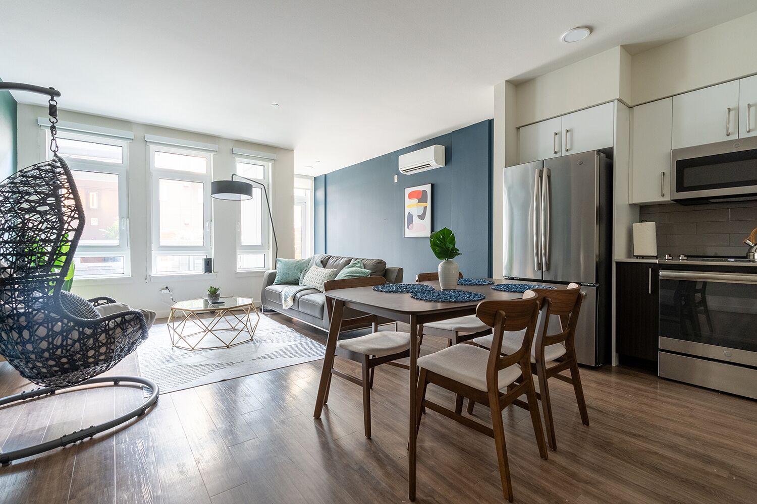 Et moderne køkken med stålemmer, et spiseområde med et træbord og stole, samt en hyggelig siddekrog med sofa og hængestol.