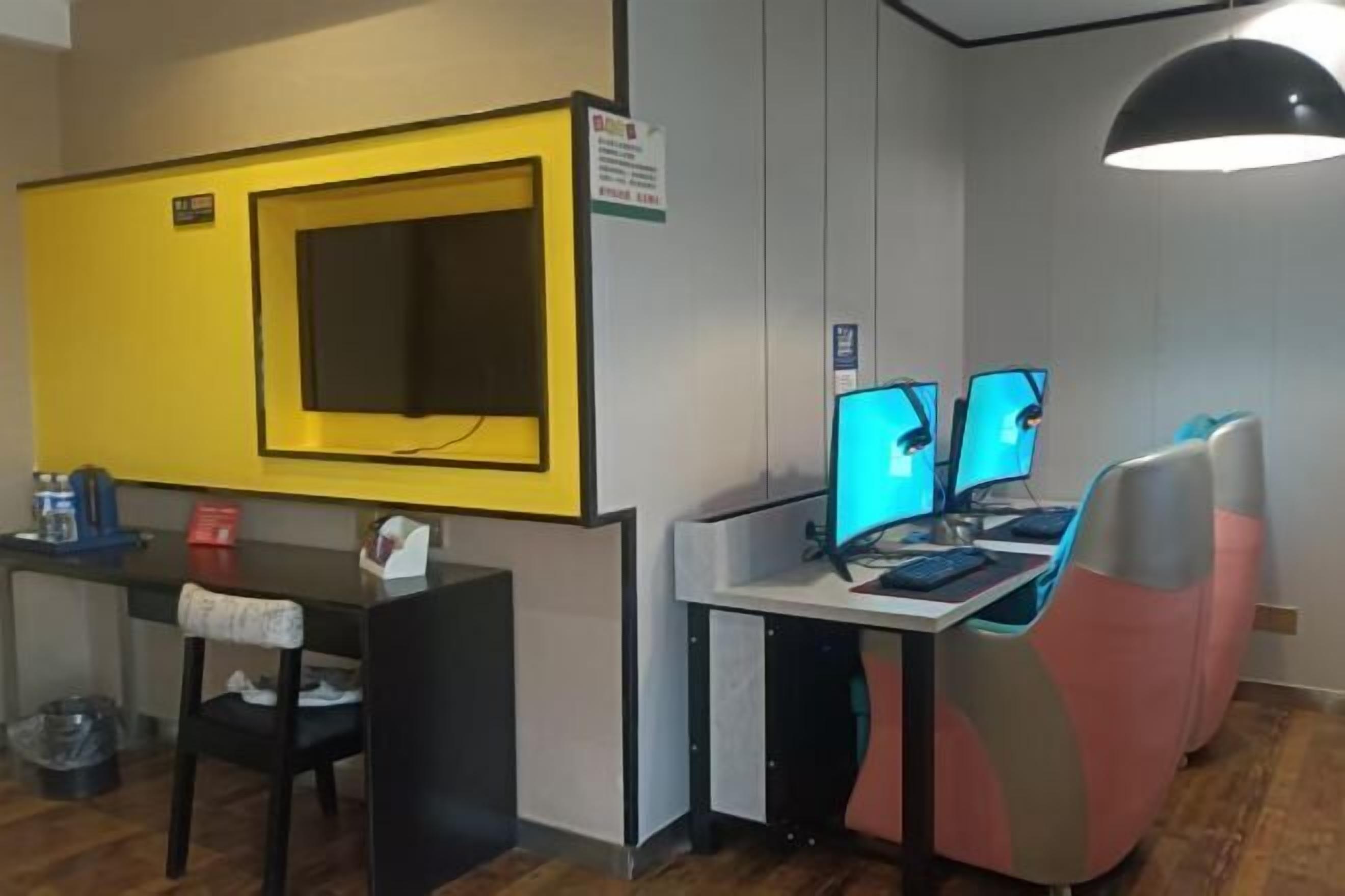 A room with a yellow accent wall, a desk with two monitors, a chair, and a hanging light fixture. - Zunyi Hengsheng Hotel (Yuqing)