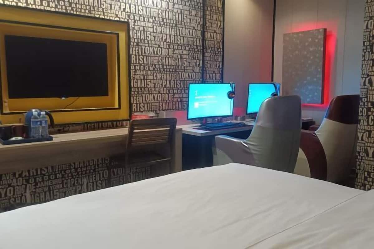 A hotel room with a large bed, two computer monitors, a desk with a chair, and a wall with city names.