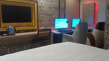 A hotel room with a large bed, two computer monitors, a desk with a chair, and a wall with city names.