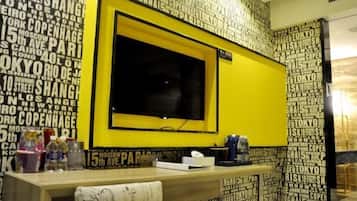 A hotel room with a large TV on a yellow stand, a wooden desk with a tissue box, and a wall with city names in black and white.