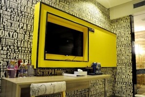A hotel room with a large TV on a yellow stand, a wooden desk with a tissue box, and a wall with city names in black and white. - Zunyi Hengsheng Hotel (Yuqing)