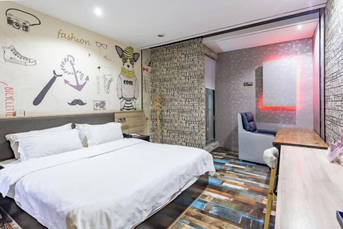 A hotel room with a bed, a desk, a chair, and a wall with various fashion-related drawings.