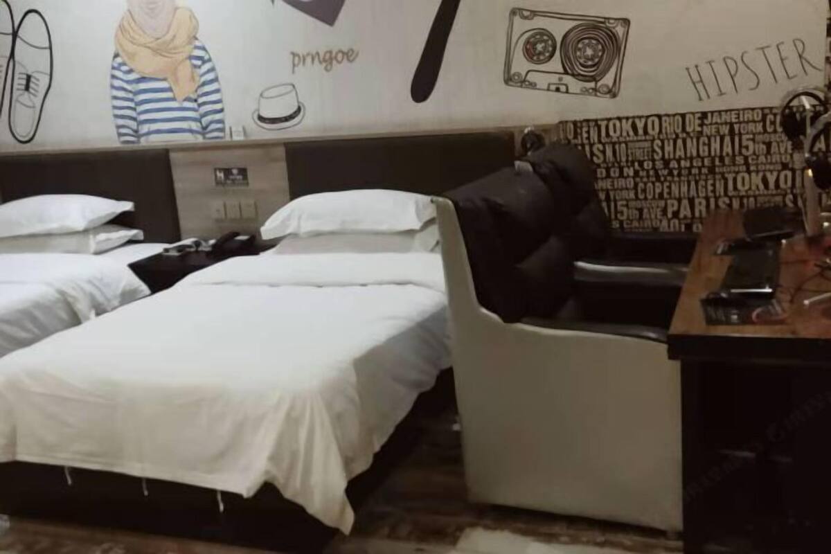 A hotel room with two beds, a desk, and a wall with various city names and a drawing of a person.