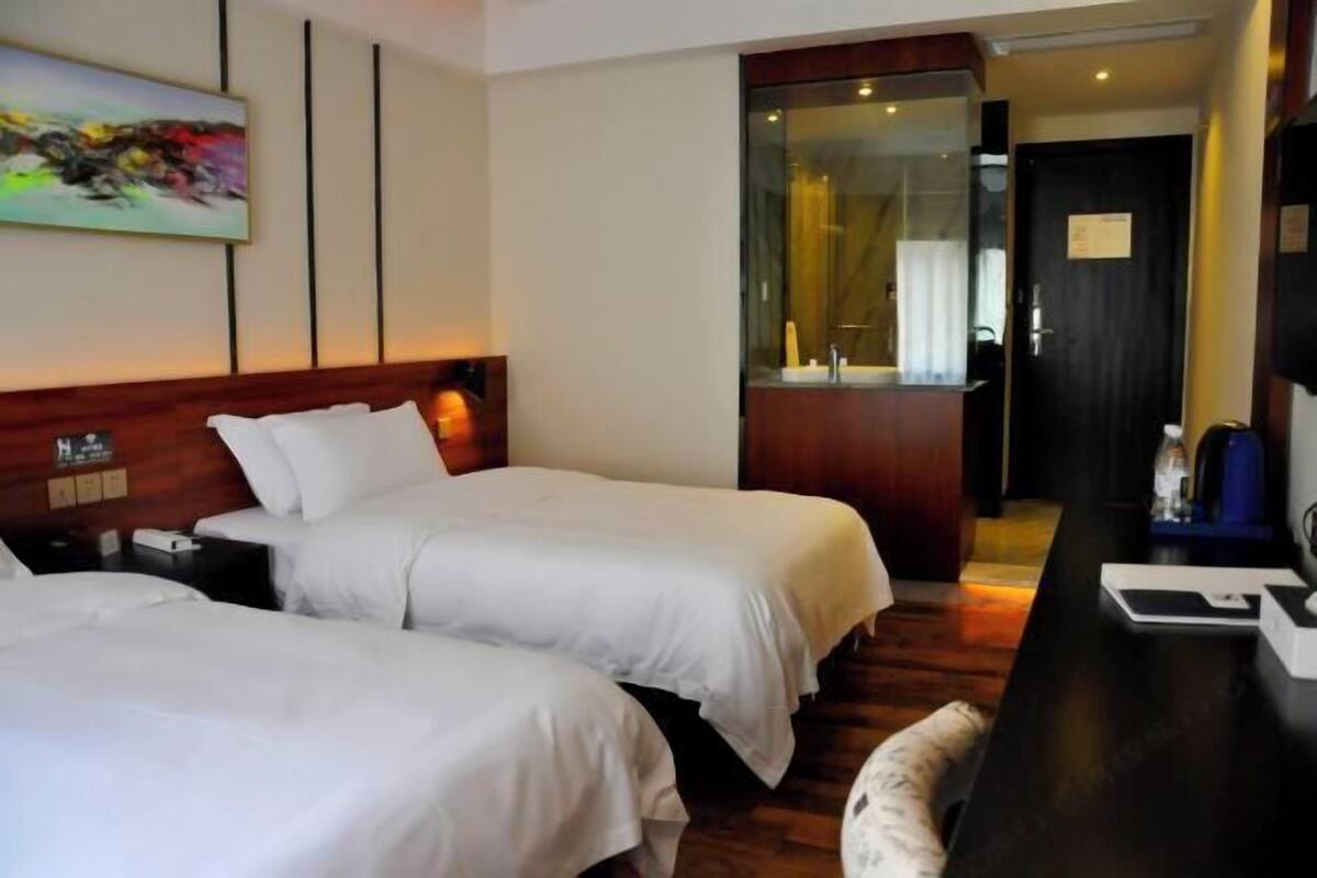 A hotel room with two single beds, a wooden headboard, a desk with a chair, and a bathroom visible through a glass door.