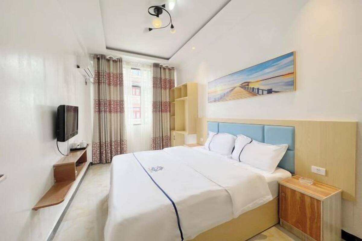 A hotel room with a bed, a TV, a painting, and a window with curtains.