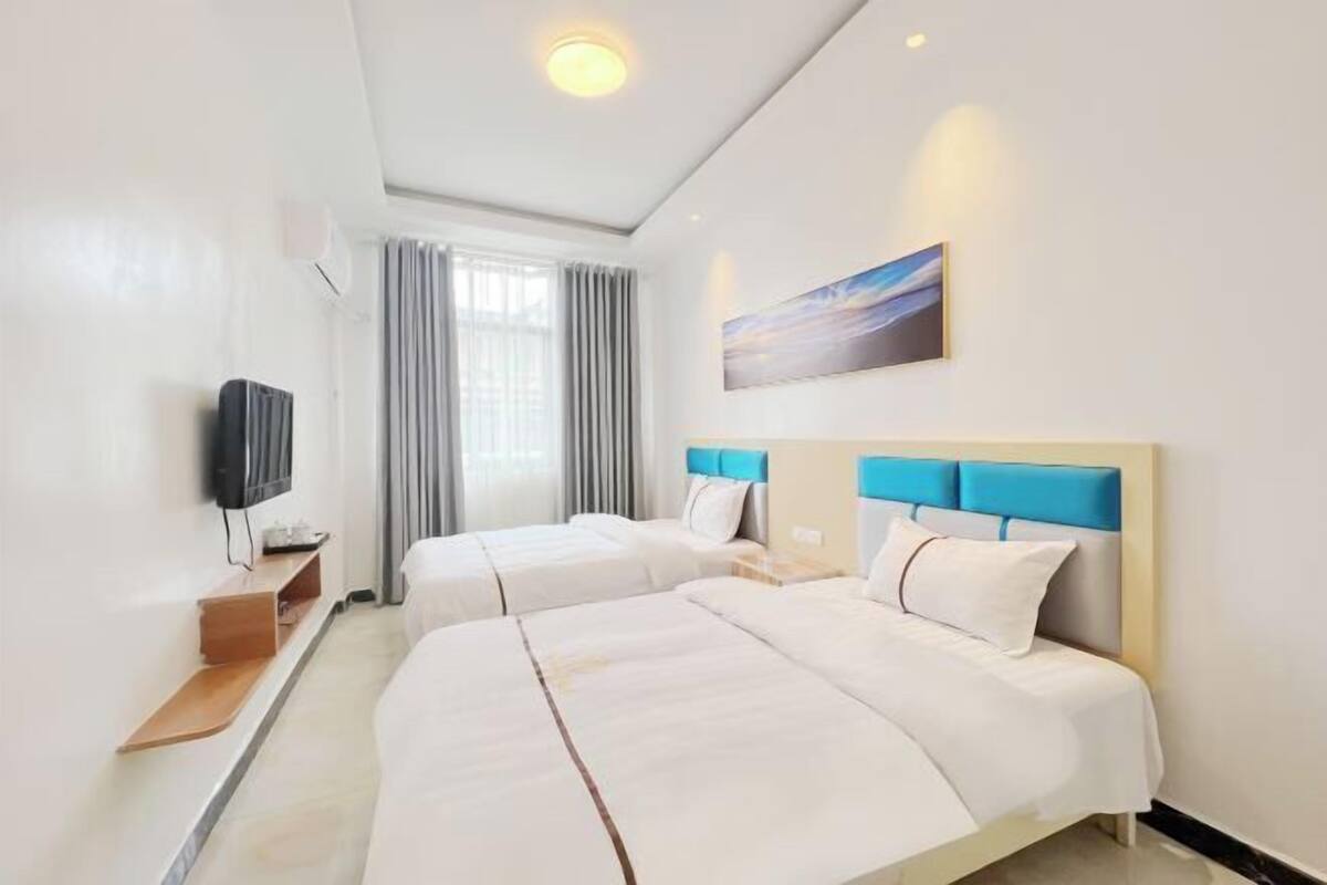 A hotel room with two beds, a TV, a painting, and a window with curtains.