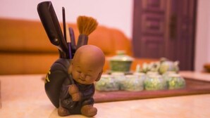 Una figurita de un monje sosteniendo un pincel, con un fondo desenfocado que incluye un sofá y un aparador con objetos decorativos.