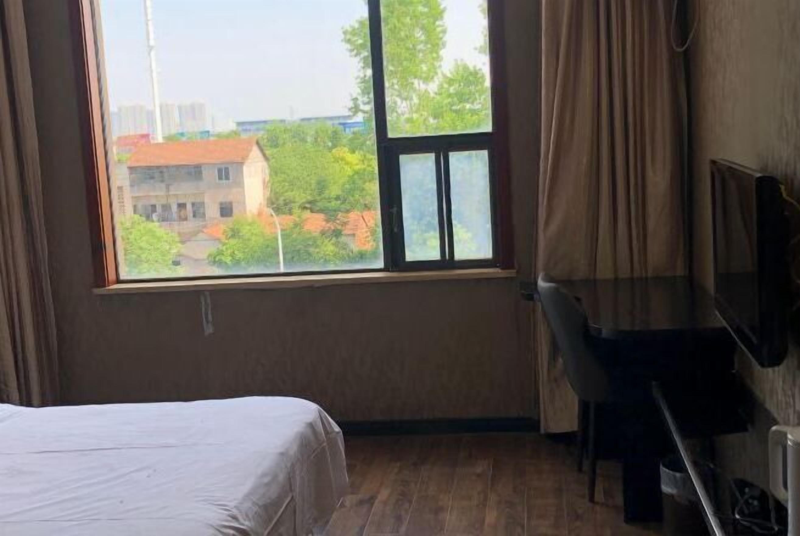 A hotel room with a bed, a desk, and a large window offering a view of buildings and trees.