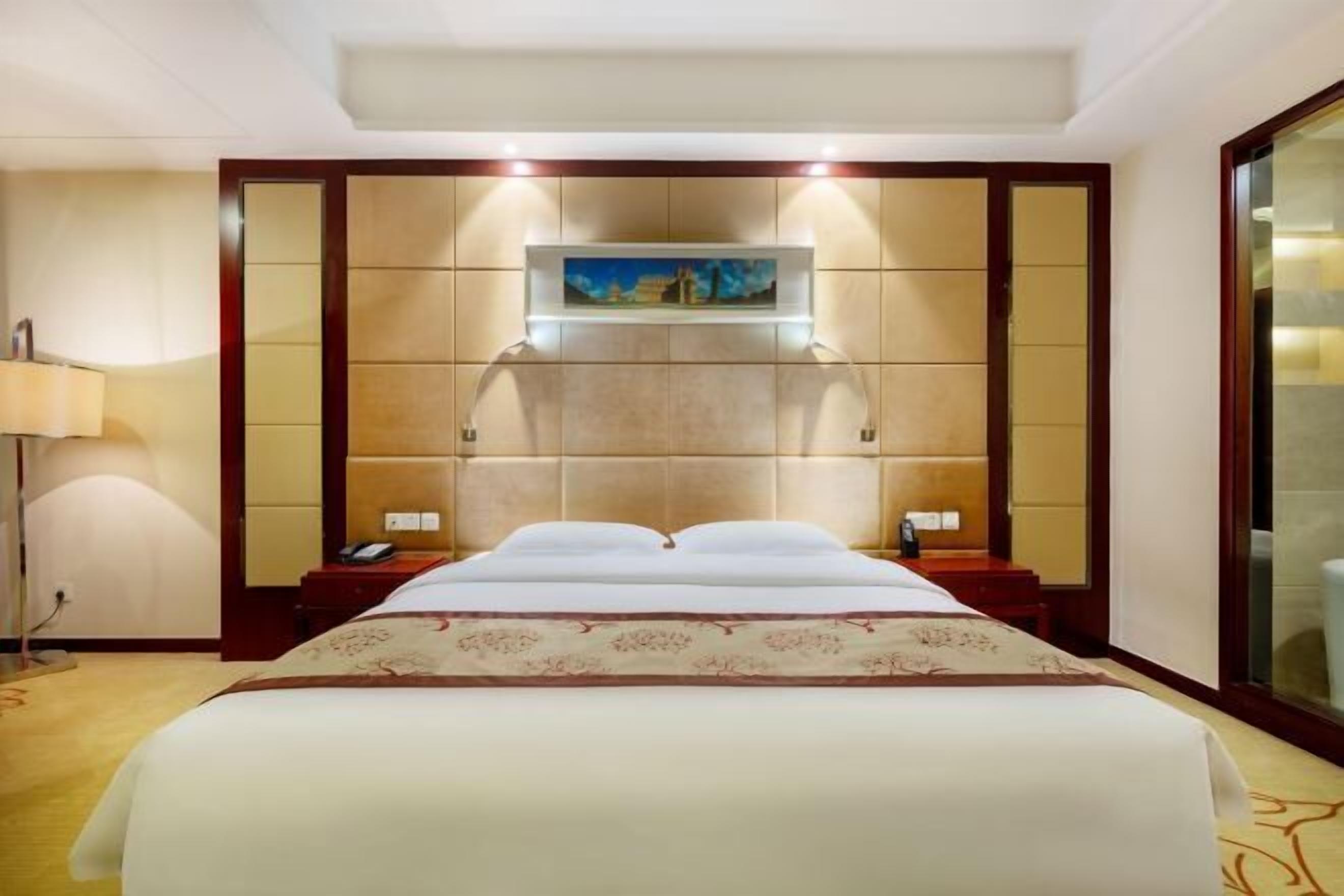 Ein Hotelzimmer mit einem großen Bett, einem Bild über dem Kopfteil, einem Nachttisch mit einer Lampe und einem großen Fenster.
