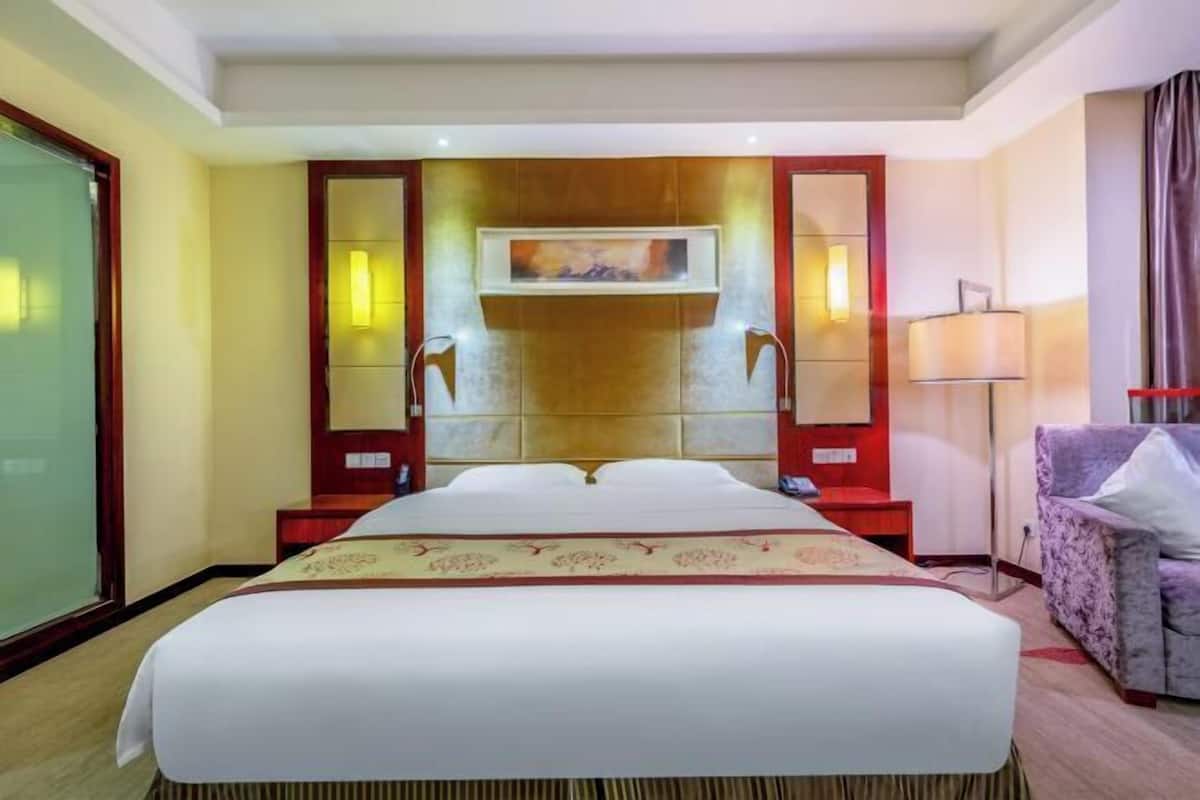 A hotel room with a large bed, a headboard with a painting, a side table, a lamp, and a chair.