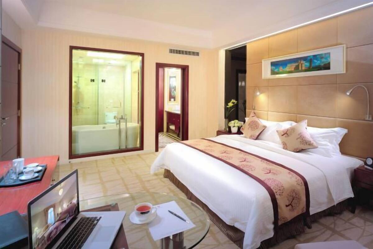 A hotel room with a bed, a laptop on a small table, a glass shower enclosure, and a painting on the wall.