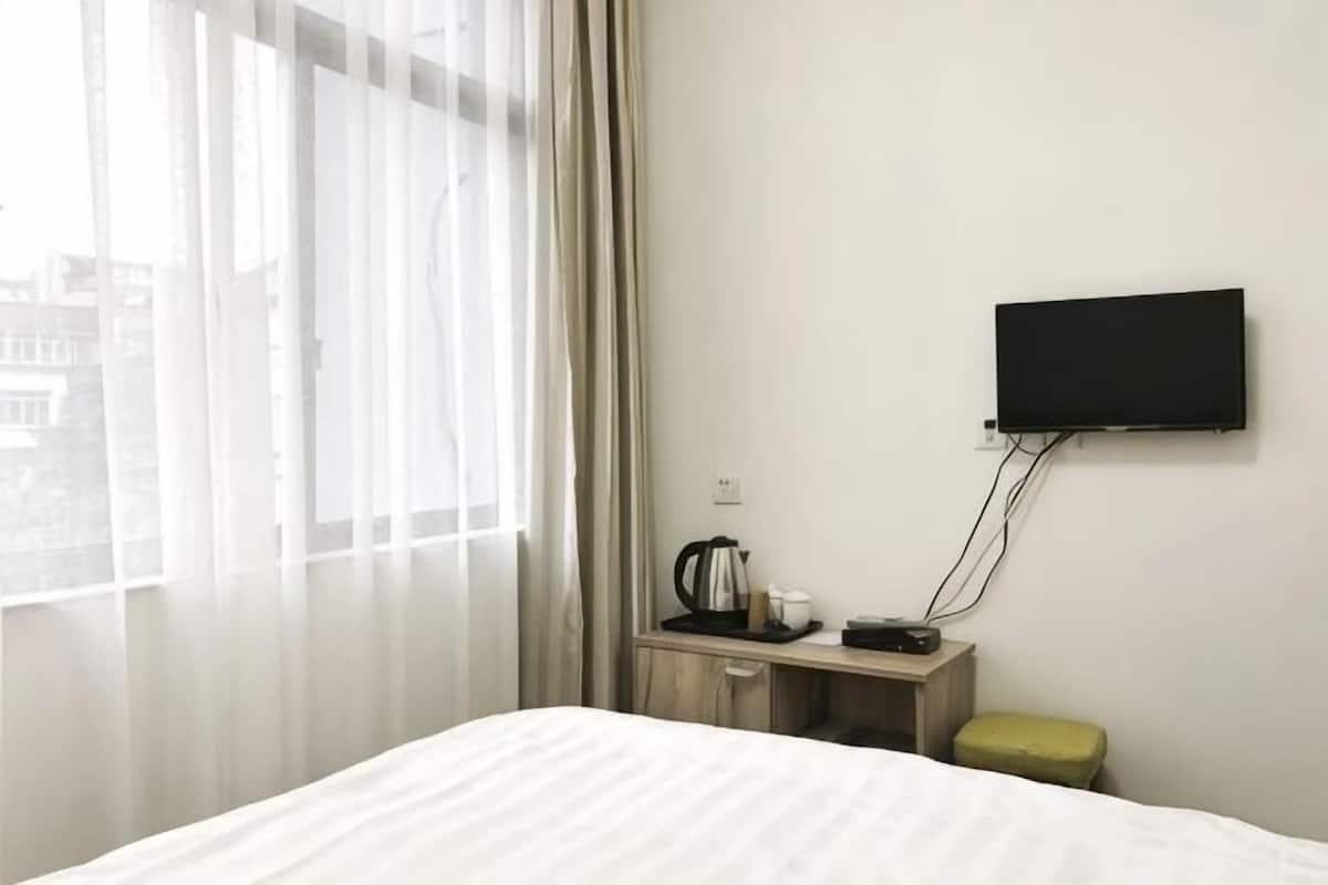 Habitación de hotel con una cama, un televisor montado en la pared, una mesita con una tetera y tazas, y una ventana con cortinas de encaje.