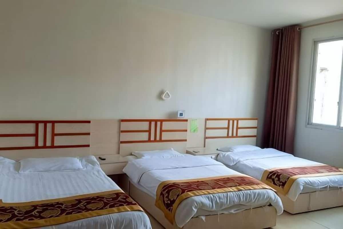 A hotel room with three beds, a window with curtains, a wall-mounted light, and a small wall clock.