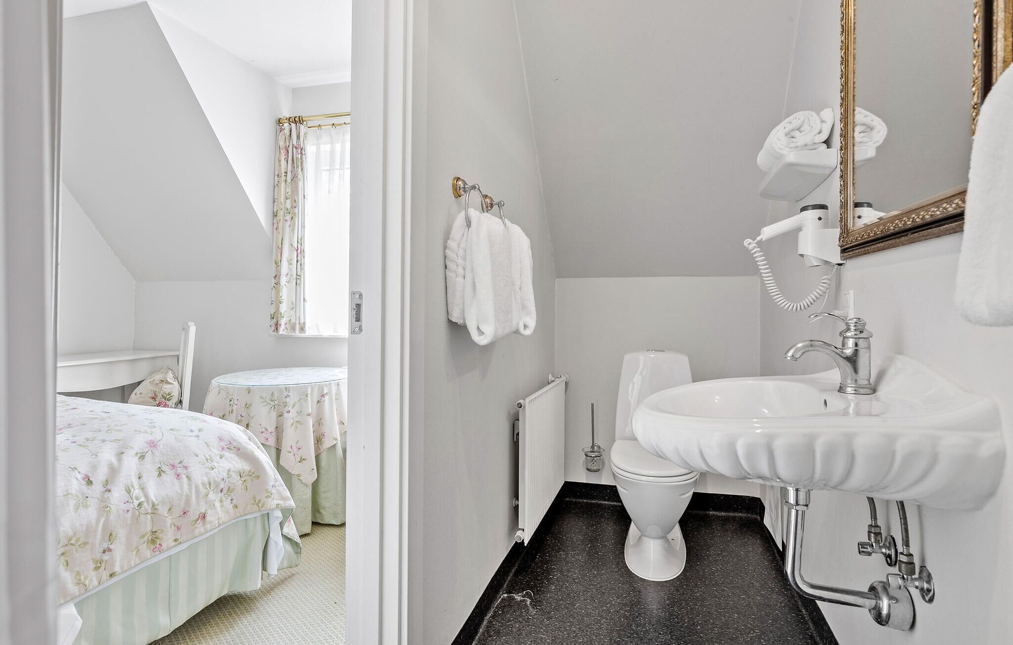 A bathroom with a white sink, a toilet, and a shower area. There is a mirror with a gold frame, and towels are hung on the wall.