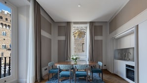 A dining area with a wooden table and blue upholstered chairs, a decorative hanging light, a view of a stone building through a window, and a kitchen with white cabinets and a built-in oven.