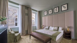 A hotel room with a large bed, a bench, a desk, and a chair. There are curtains, a window, and framed pictures on the wall.