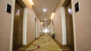 Een lange, smalle hotelgang met een vloerkleed met patroon, lichte muren en een rij deuren aan één kant.