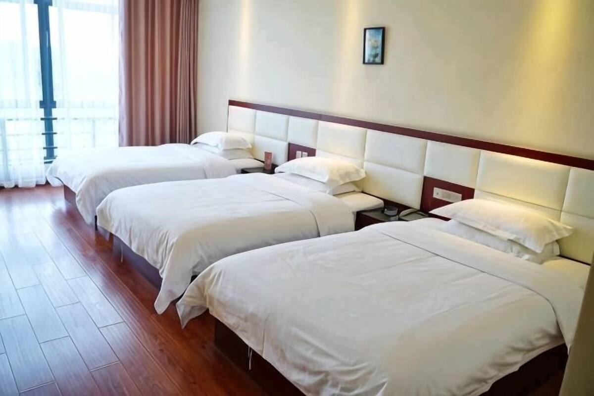 Een hotelkamer met drie bedden, houten vloeren en een groot raam met gordijnen.