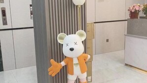 Een beer mascotte in een lobby met een verticaal gestreepte muur, een receptiebalie en een bloemstuk.