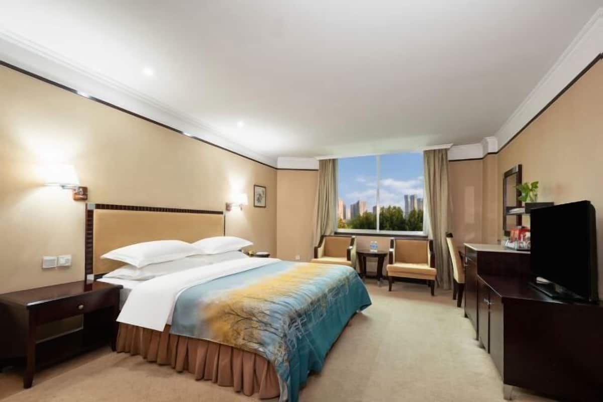 Ein Hotelzimmer mit Bett, Schreibtisch, Sessel, Fernseher und einem Fenster mit Blick auf die Stadt.