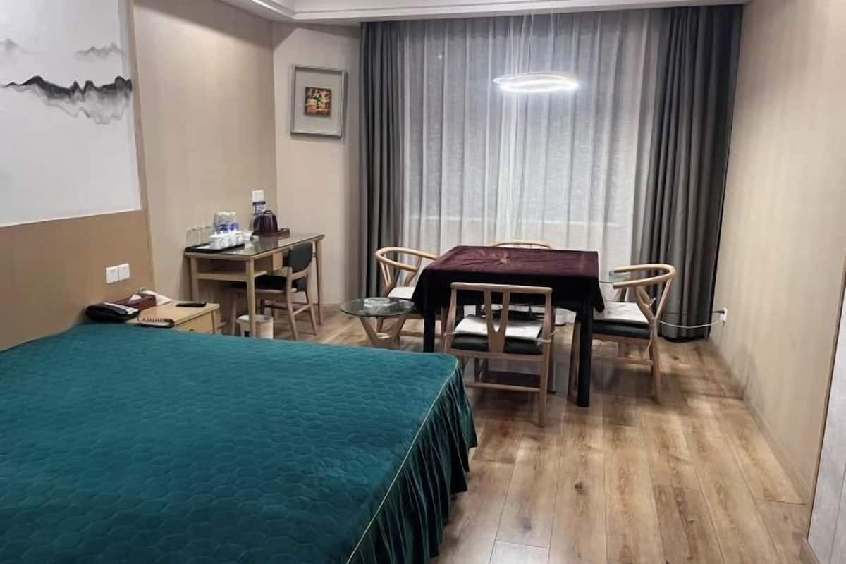 Ein Hotelzimmer mit einem Bett, einem Schreibtisch mit Stuhl, einem Tisch mit Stühlen, einem Fenster mit Vorhängen und einem Bild an der Wand.