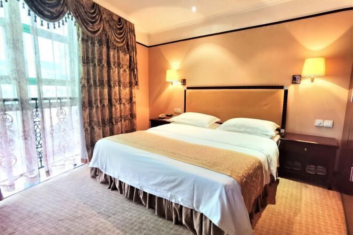 Ein Hotelzimmer mit einem großen Bett, zwei Nachttischlampen, einem Nachttisch, einem Fenster mit Vorhängen und Blick nach draußen.