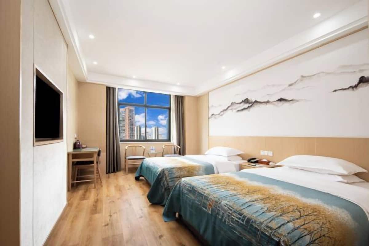 Ein Hotelzimmer mit zwei Betten, einem Schreibtisch und einem großen Fenster mit Blick auf die Stadt.