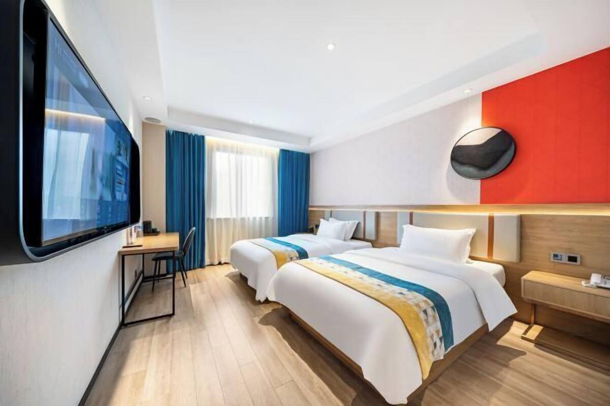 Hotelkamer met twee bedden, een bureau, een stoel, een aan de muur bevestigde televisie en een groot raam met gordijnen.