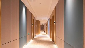 Un pasillo largo y estrecho con piso y paredes de color claro, bordeado por puertas distribuidas regularmente e iluminado por luces del techo.