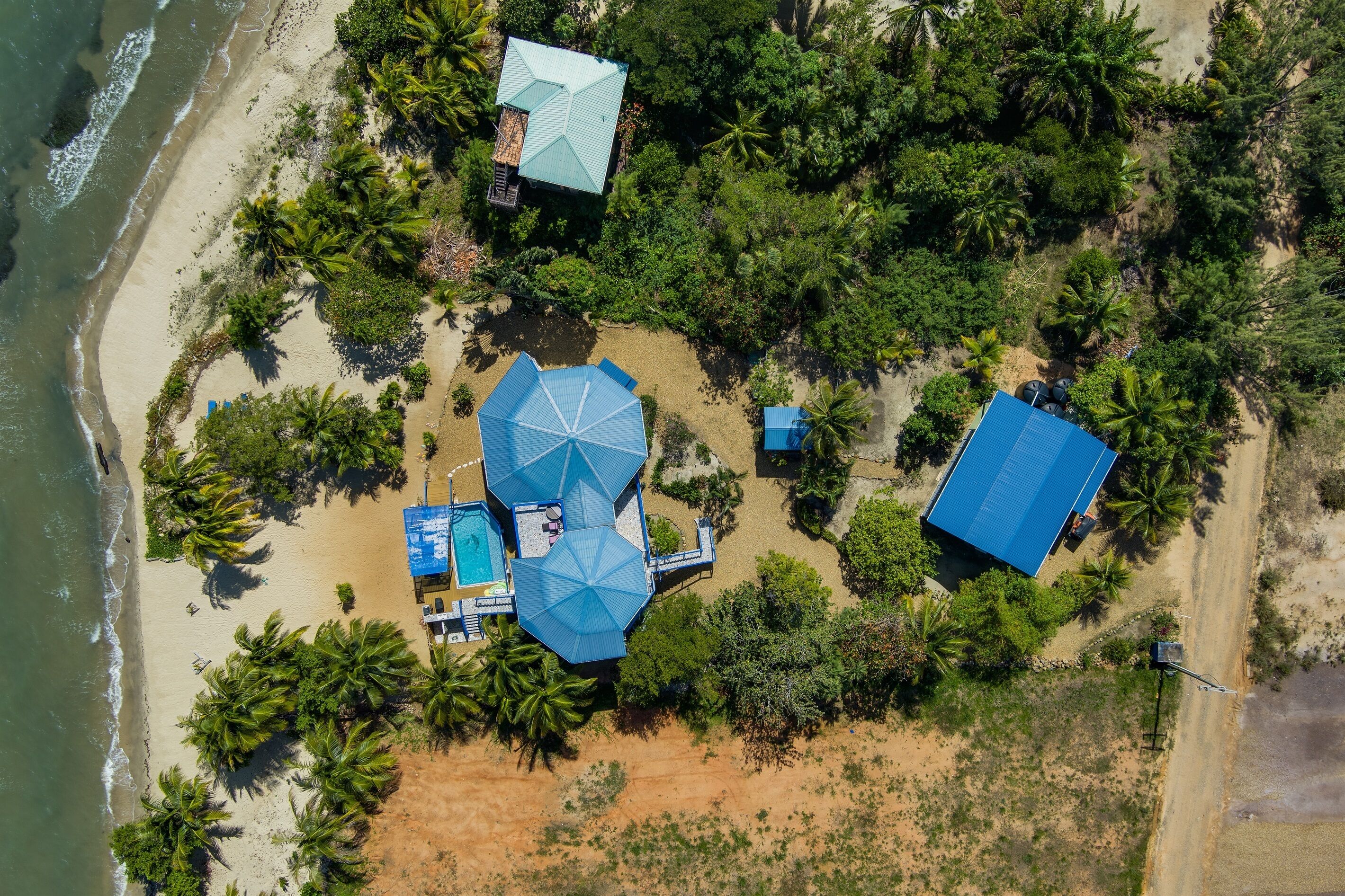 An aerial view of a beachside property with a swimming pool, umbrellas, and a large blue-roofed building.
