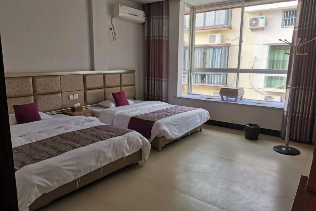 A hotel room with two beds, a window with a view of buildings, an air conditioning unit, and a small table.