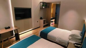 Ein Hotelzimmer mit zwei Betten, einem Schreibtisch, einem an der Wand befestigten Fernseher und einem kleinen Regal mit einer Pflanze.