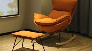 Ein Zimmer mit Schaukelstuhl, Hocker, Projektionsschirm und einem mit Vorhängen versehenen Fenster.