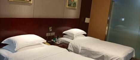 A hotel room with two single beds, a wooden headboard, a small table with a lamp, and two paintings on the wall.