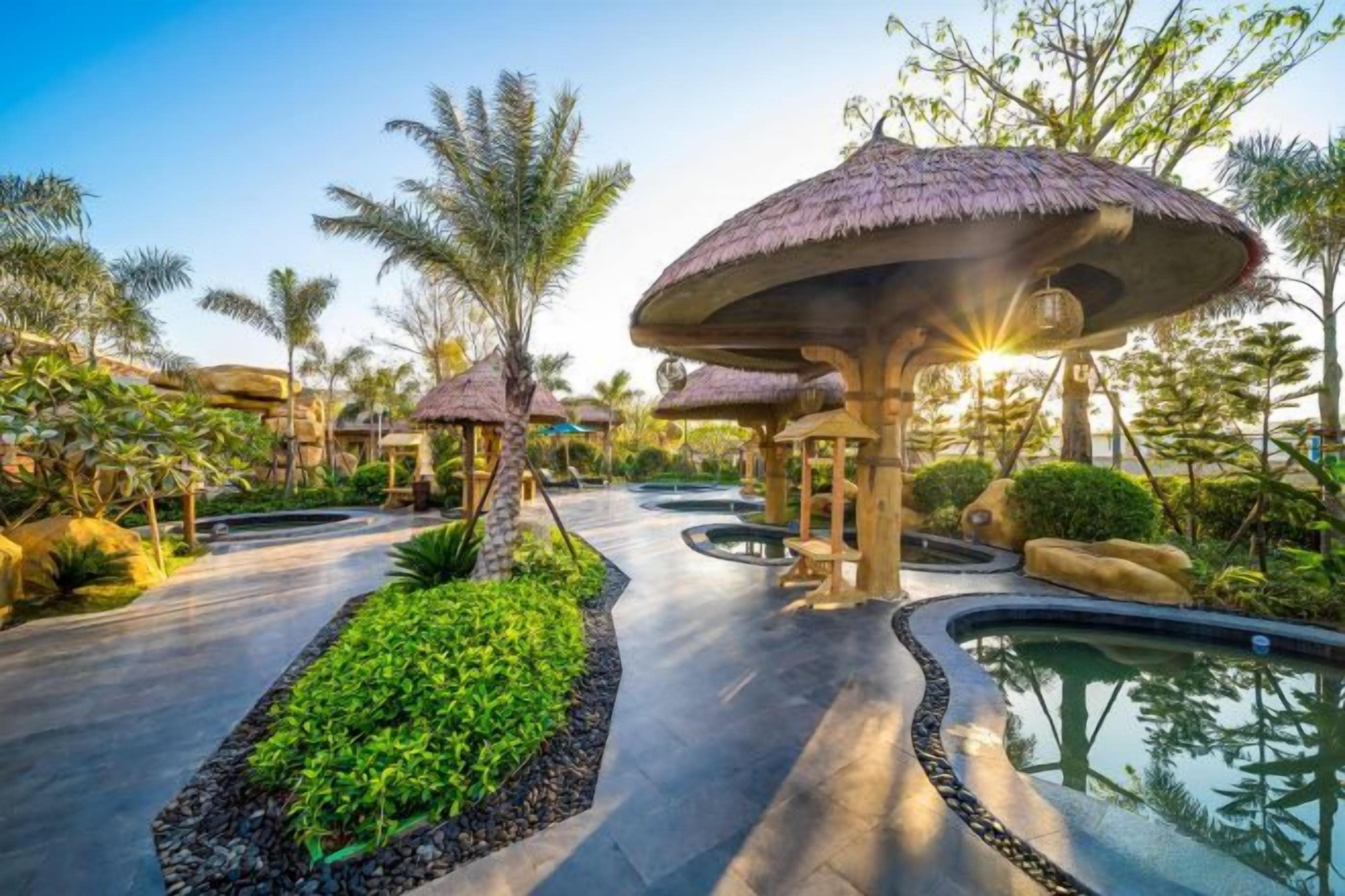 A resort pathway with palm trees, a water feature, and thatched-roof structures.