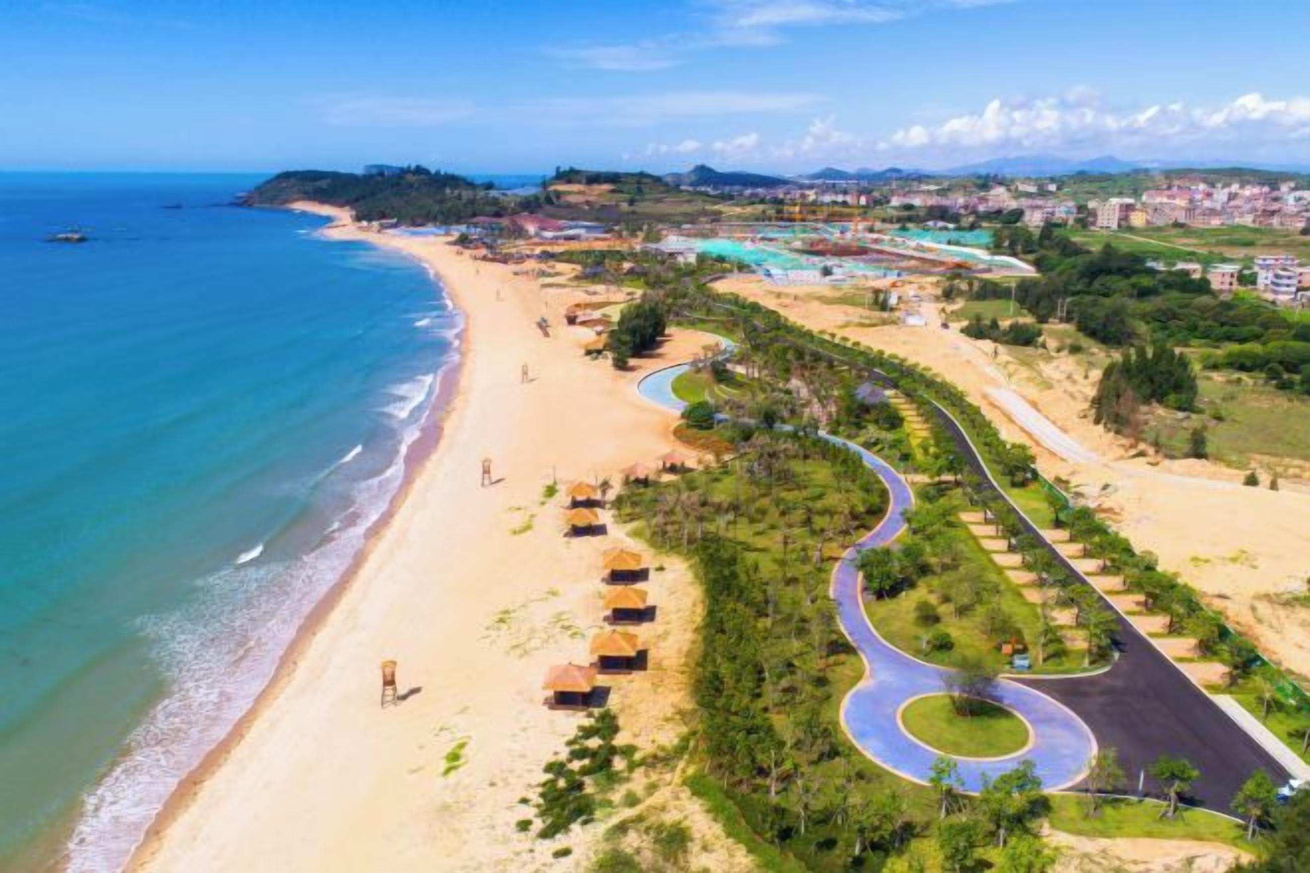 Una carretera costera con una playa a un lado y un área ajardinada con árboles y una carretera curva al otro.