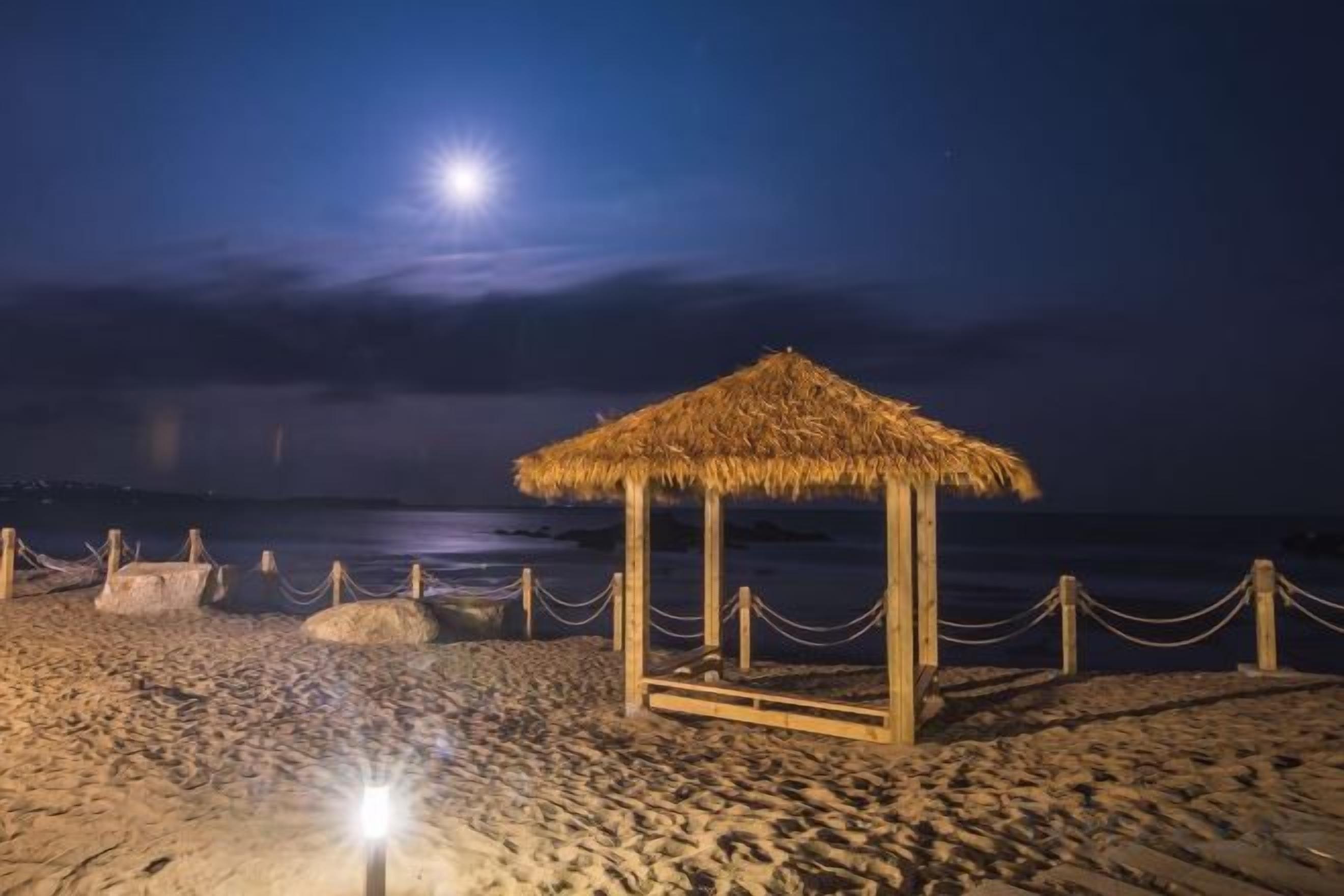 Uma praia com uma estrutura de telhado de palha, cerca de arame e um corpo d'água sob um céu noturno com a lua visível.