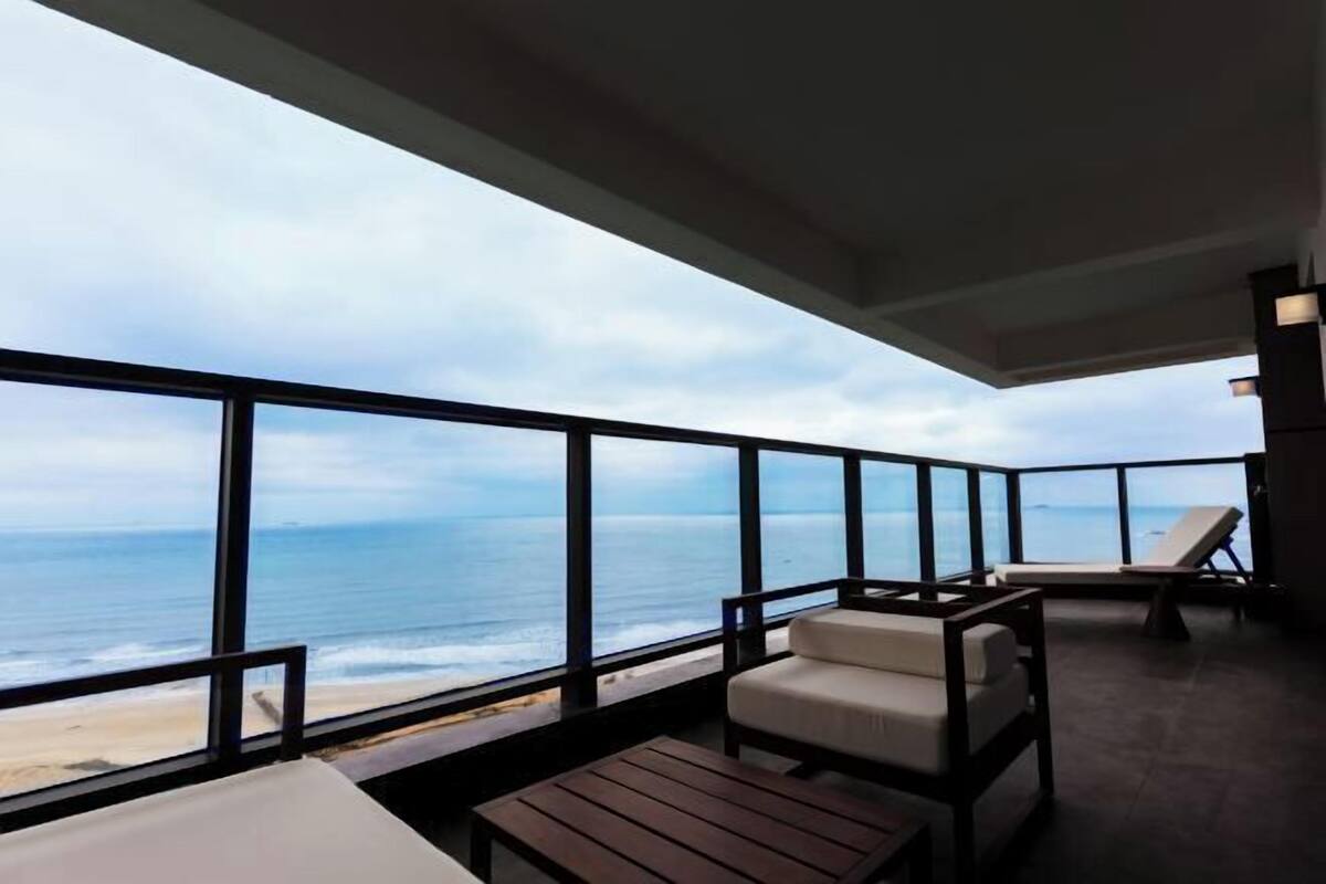 Un balcon avec vue sur l’océan, deux chaises blanches avec coussins et une table en bois.