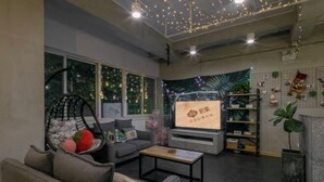 Een moderne woonkamer met een grijze bank, een houten salontafel en een televisiescherm waarop een poster staat. Er zijn grote ramen met gordijnen, een plant en een plank met decoratieve objecten.