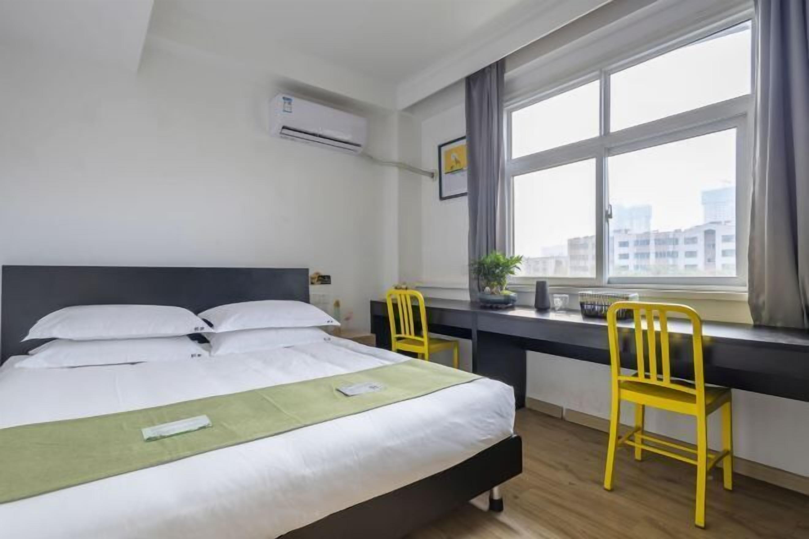 A hotel room with a bed, a desk, two chairs, an air conditioner, a window with a view of buildings, and a potted plant.