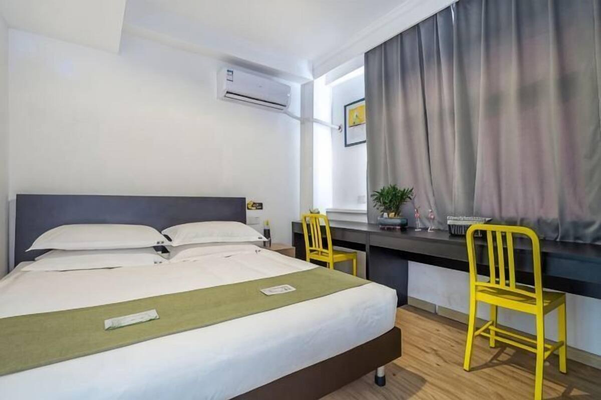 A hotel room with a bed, a desk, two chairs, a window with curtains, and an air conditioning unit.