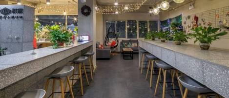 Una zona de bar con varios taburetes y una barra larga, decorada con plantas y guirnaldas de luces.