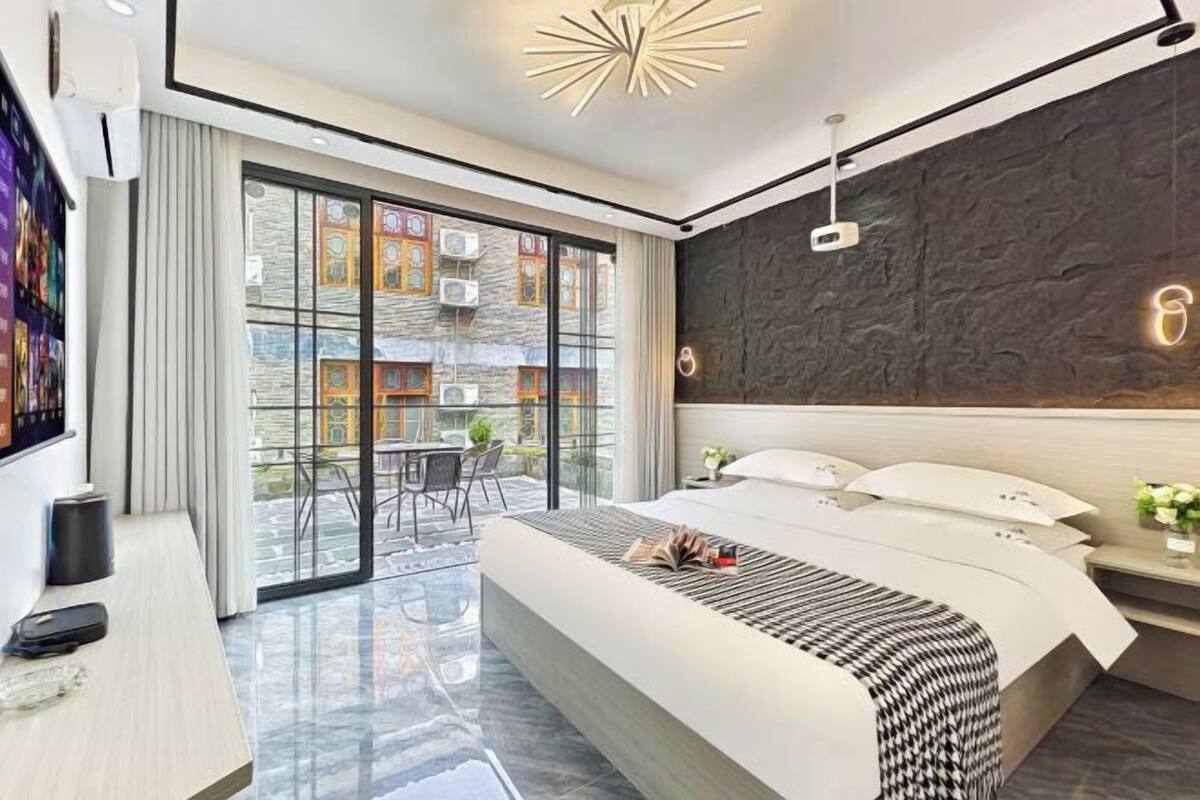 A modern hotel room with a large bed, a desk, a TV, and a balcony with a view of buildings.