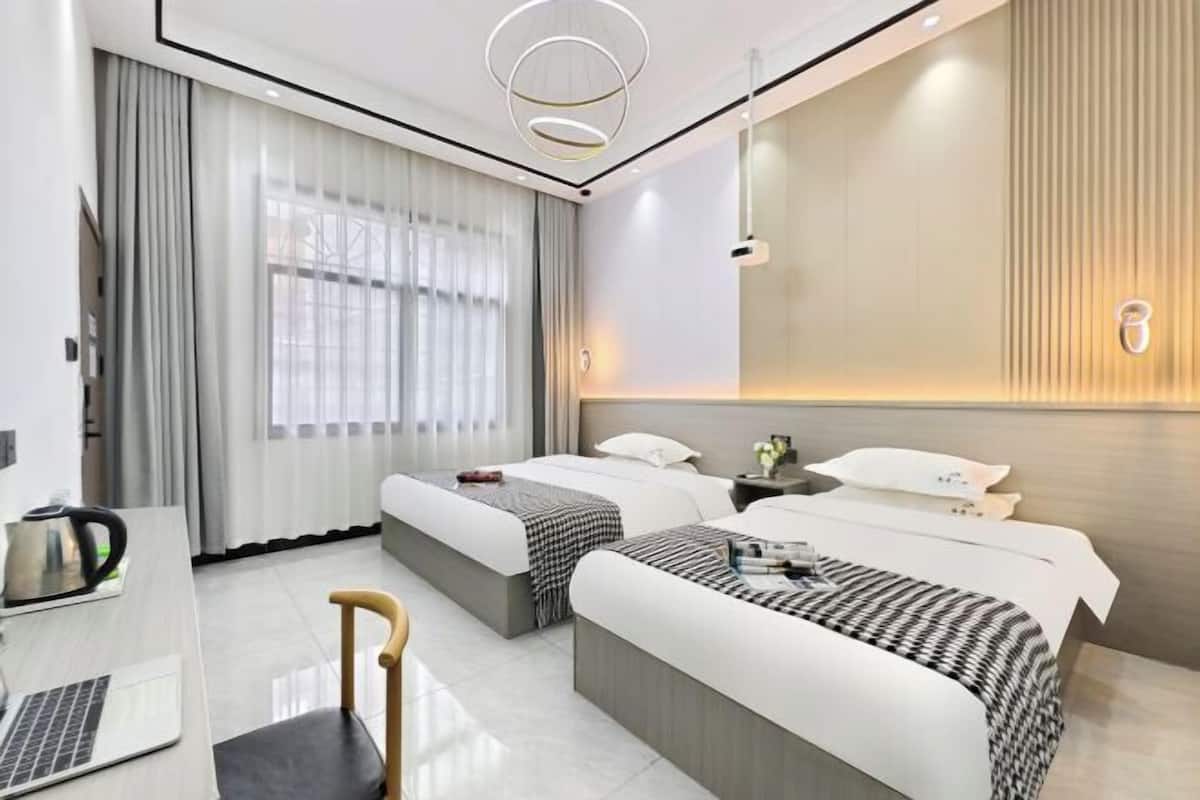 A hotel room with two beds, a desk with a laptop, a chair, and a large window with curtains.