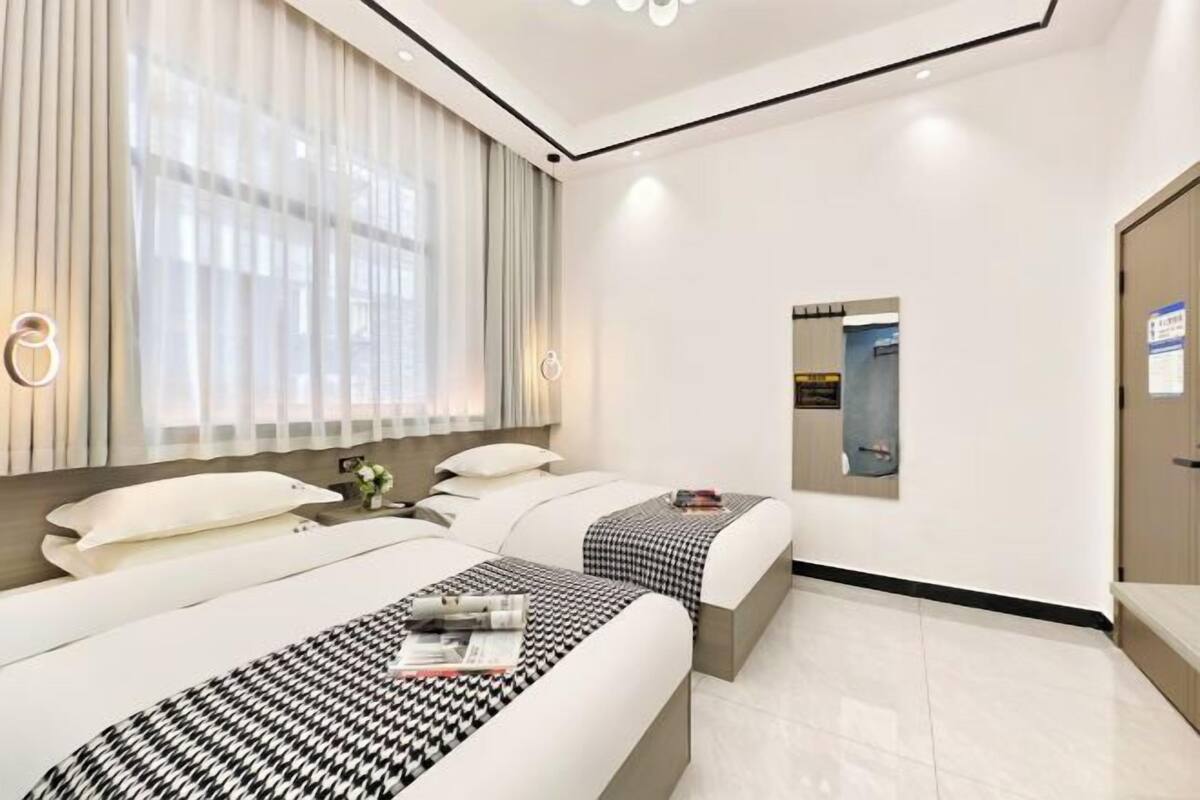 A hotel room with two beds, a large window with sheer curtains, a mirror, and a door with a sign.