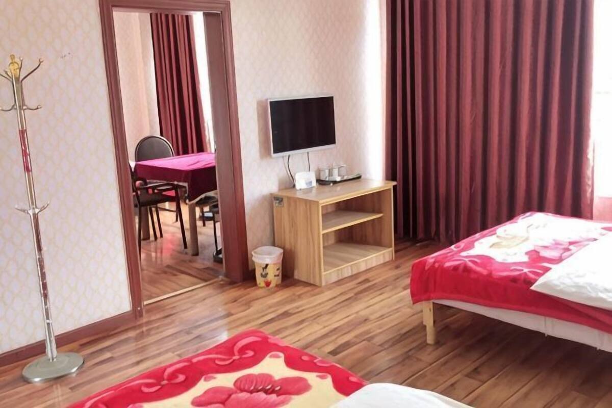 A hotel room with a wooden floor, a bed with a red and white bedspread, a desk with a TV, a coat rack, and a dining area visible through an open door.