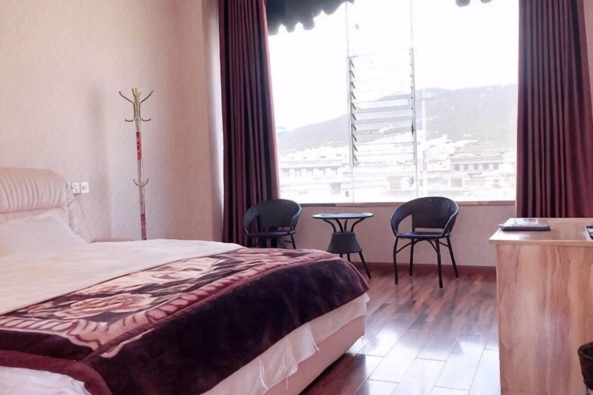 A hotel room with a bed, a small table, two chairs, a desk, and a view of buildings through large windows.