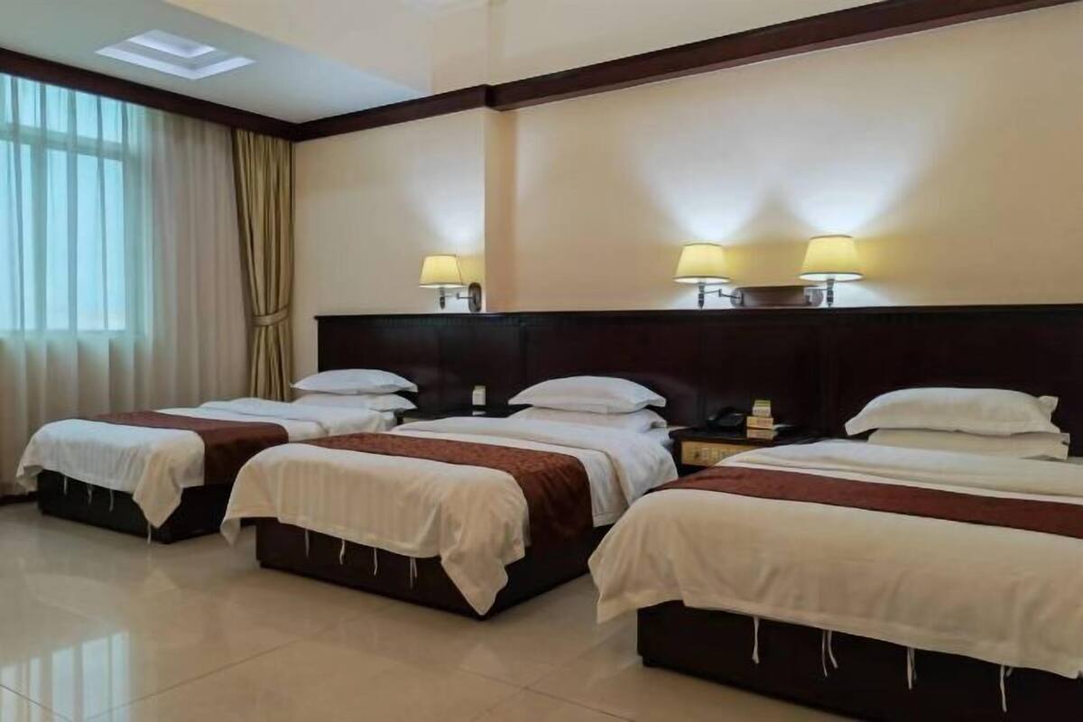A hotel room with three beds, a large window with curtains, and wall-mounted lamps.