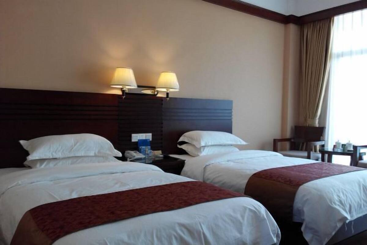A hotel room with two beds, a nightstand with a lamp, a window with curtains, and a small table with a chair.